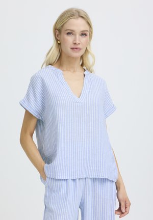 Vaaleatukkainen nainen, joka pukeutunut vaaleansinivalkoisiin pystysuoriin raidoitettuihin lyhythihaiseen paitaan ja vastaaviin housuihin, seisoo käsi taskussa.