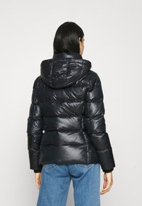 Kvinna som bär en svart quiltad pufferjacka med huva, stående med ryggen mot kameran, i blå jeans mot en plain ljus bakgrund.
