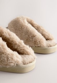 Chaussons beiges et moelleux à design ouvert, dotés d'une fausse fourrure douce et d'une semelle extérieure texturée. Le nom de la marque "NEXT" est embossé sur le côté.