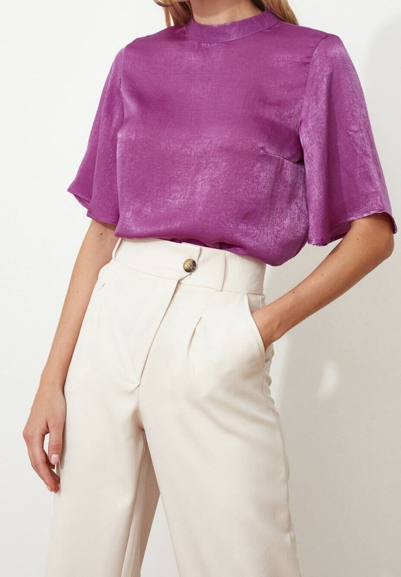 Top di raso viola con maniche corte e colletto alto, abbinato a pantaloni beige a vita alta con bottoni decorativi e pieghe.