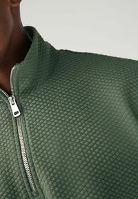 Veste zippée vert kaki mettant en valeur un tissu texturé à motif en losange et un zip argenté au niveau du cou, soulignant un design moderne.