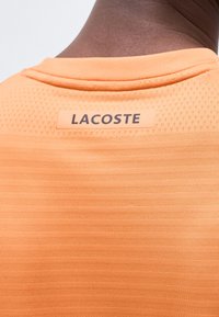 Κοντινό πλάνο πορτοκαλί υφασμάτινου αθλητικού πουκαμίσου στην πλάτη ενός ατόμου με την ετικέτα "LACOSTE" κάτω από το γιακά του λαιμού.