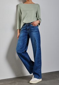 Person som bär lösa blå jeans, ljusgrön långärmad tröja, vita sneakers, står med ena handen i fickan mot en neutral vägg.