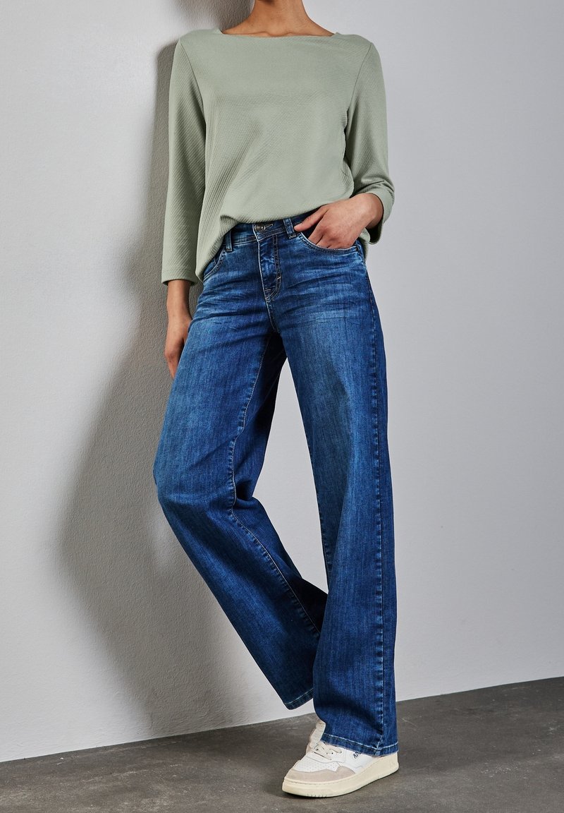 Person som bär lösa blå jeans, ljusgrön långärmad tröja, vita sneakers, står med ena handen i fickan mot en neutral vägg.