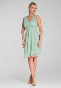 Mintgrünes Kleid mit V-Ausschnitt und ärmellos Design. Der knielange Rock hat eine geschichtete, fließende Textur. Einfache Sandalen mit Riemen.