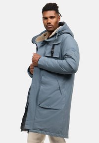Kék, hosszú kabát, kapucnival, texturált bolyhos béléssel, elülső zsebekkel és cipzáras záródással. Fekete fém részletekkel.