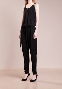 Top negro sin mangas con tirantes, combinado con pantalones negros ajustados. Cuenta con una cintura elástica y acentos de metal dorado. Llevado con tacones altos negros.