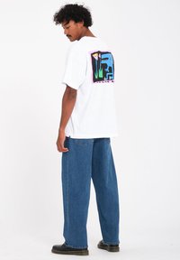 White oversized t-shirt with a colorful graphic on the back, featuring a blue creature and plants, paired with loose blue jeans.