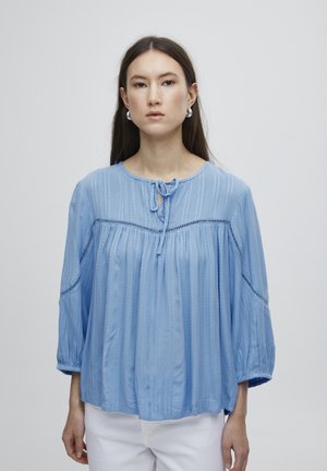 Helle blaue, langärmlige Bluse aus weichem Stoff. Sie hat einen runden Halsausschnitt mit einer Schleife, vertikale Falten und Aussparungen an den Ärmeln.