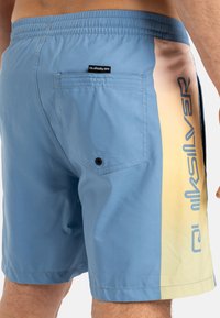 Világoskék fürdőnadrág, amelynek oldalán puha sárga és rózsaszín átmenetes panel található, hátul zsebes és Quiksilver logóval ellátott. Sima textúra.
