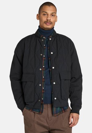 Quilted black bomber jacket with snap buttons, two front pockets, ribbed cuffs, layered over a green plaid flannel shirt. Textured fabric.