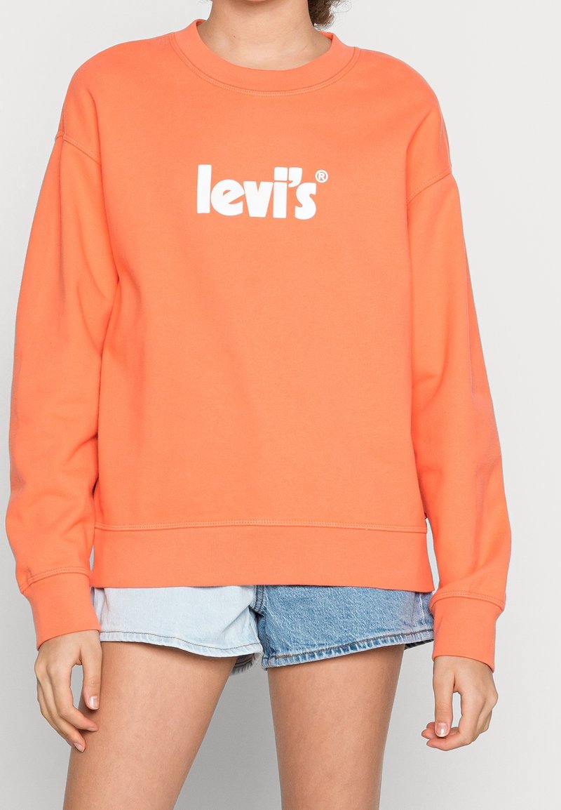 Persona con sudadera naranja de Levi's y pantalones cortos de mezclilla bicolor, de pie contra un fondo claro y liso.