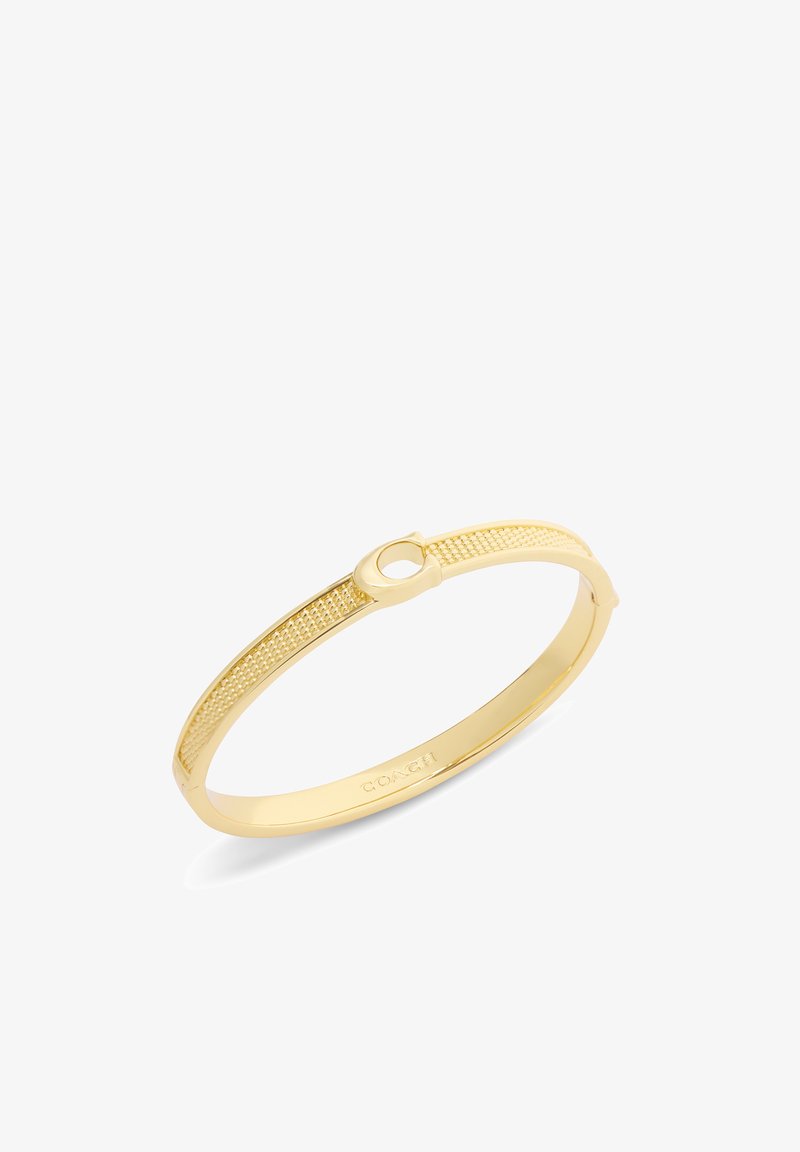 Bracelet jonc doré avec détails texturés et fermoir circulaire, gravé du nom de la marque « Coach » à l'intérieur.