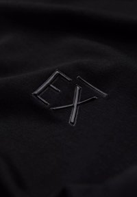 Czarny materiał z wypukłym czarnym logo "EX" w błyszczącym wykończeniu. Tekstura jest gładka z subtelnym połyskiem, podkreślająca monochromatyczny design.