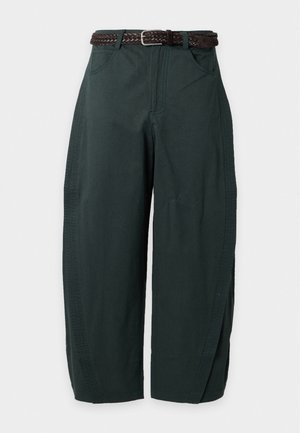 Mujer de pie con las manos en los bolsillos, vestida con una camisa polo de punto blanca a rayas, pantalones anchos verde oscuro, cinturón negro y zapatos planos negros.