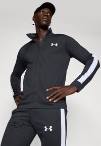 Fekete sportos zipzáras dzseki fehér oldalsávokkal és Under Armour logóval. Párosítva fekete nadrággal, fehér részletekkel.