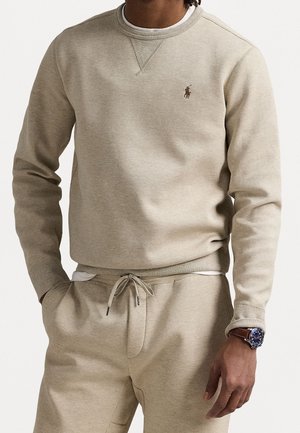Mand iført beige sweatshirt og matchende snøre bukser med hænderne i lommerne og et armbåndsur med brun læderrem.
