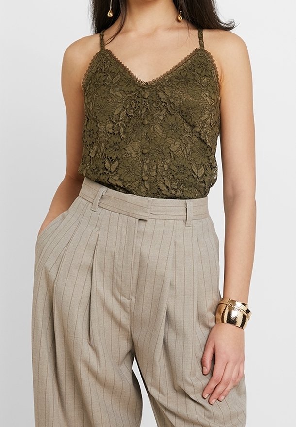 Femme portant un caraco en dentelle vert olive avec des boutons, associé à un pantalon plissé beige à fines rayures taille haute et un bracelet manchette en or.