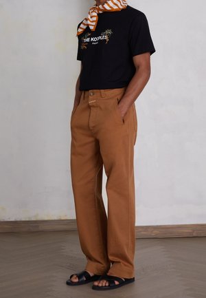 Mężczyzna ubrany w czarną koszulkę z nadrukiem, brązowe proste spodnie oraz czarne sandały, z pomarańczowo-białym paskowanym szalikiem związanym wokół szyi.