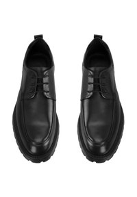 Une paire de chaussures de ville en cuir noir à lacets avec de semelles en caoutchouc épaisses, vue d'en haut sur un fond blanc.