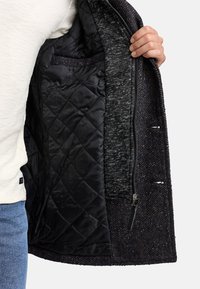 Interior negro acolchado de una chaqueta de tweed oscuro, con un bolsillo lateral y una tela de textura contrastante en el borde. Cierre con cremallera.