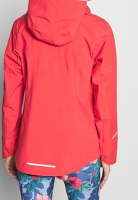 Rød vandtæt jakke med hætte, letvægtsstof, lange ærmer og reflekterende detaljer. Sættes sammen med leggings med blomstermønster.