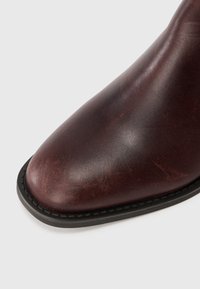 Botte en cuir brun foncé avec un bout carré arrondi et une semelle noire cousue sur fond blanc uni.