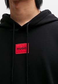 Persona vestida con una sudadera negra con cordones negros y un parche rojo etiquetado como "HUGO" en el pecho, mostrando el cuello y parte de la cara.