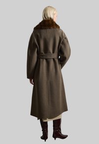 Cappotto lungo in lana marrone scuro con colletto in pelliccia marrone, vita con cintura e maniche larghe. Lunghezza gonna con cucitura dritta sul retro.