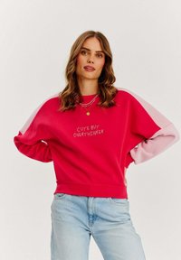 Červená mikina s růžovými rukávy, s n�ápisem "CUTE BUT OVERTHINKER" v bílé barvě. Měkká látka, uvolněný střih, design s kulatým výstřihem.