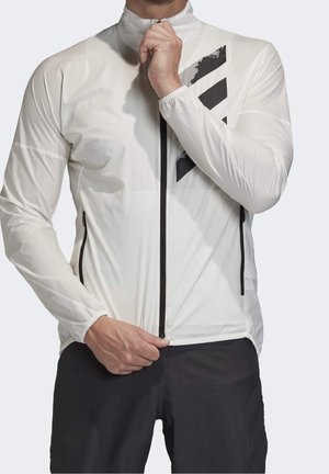 Mann, der eine weiße Jacke mit schwarzem Diagonalstreifen und Reißverschluss zumacht, dazu trägt er schwarze Hosen, vor einem schlichten Hintergrund.