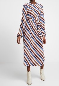 Robe midi rayée aux nuances de bleu, rose et orange ; manches longues, taille élastique et détail noué. Portée avec des bottines crème à la cheville.