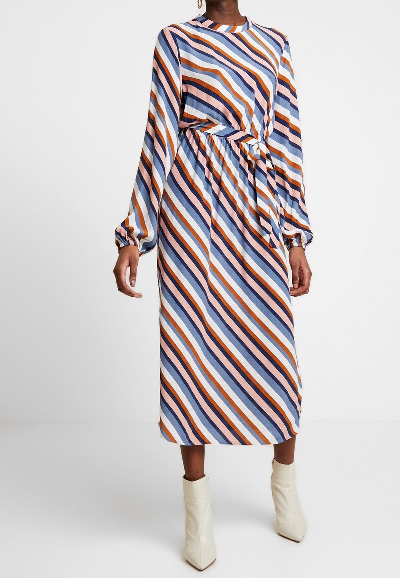 Robe midi rayée aux nuances de bleu, rose et orange ; manches longues, taille élastique et détail noué. Portée avec des bottines crème à la cheville.