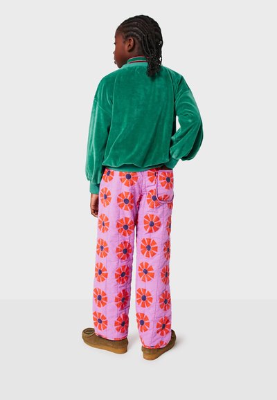 Grüne Samtjacke mit langen Ärmeln, kombiniert mit pinken gepolsterten Hosen mit roten Blumen und schwarzen Mittelpunkten sowie braunen Schuhen.