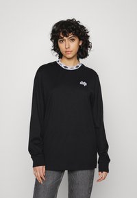 Camisa de mangas longas preta com decote redondo e logótipo gráfico em branco no peito. Por baixo, um colarinho estampado em preto e branco.