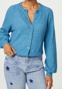 Jasnoniebieski sweter z dzianiny o teksturowanym wzorze, złote guziki w kształcie serca i luźne rękawy w kształcie balonu, noszony na dżinsach z haftem w gwiazdy.