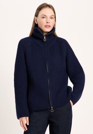 Frau trägt einen dunkelblauen, gerippten Strickpullover mit Reißverschluss und hohem Kragen, steht mit einer Hand in der Tasche vor einem schlichten Hintergrund.