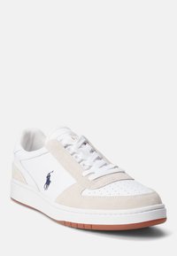 Vita sneakers med beige mocka-accenter, perforerad tå, vita snören och en brun gummisula. Har en marinblå logotyp på sidan.