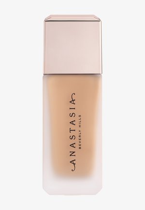 Mat glazen flesje met beige vloeibare foundation met bleekroze dop, gelabeld met “Anastasia Beverly Hills” in zwarte verticale tekst.