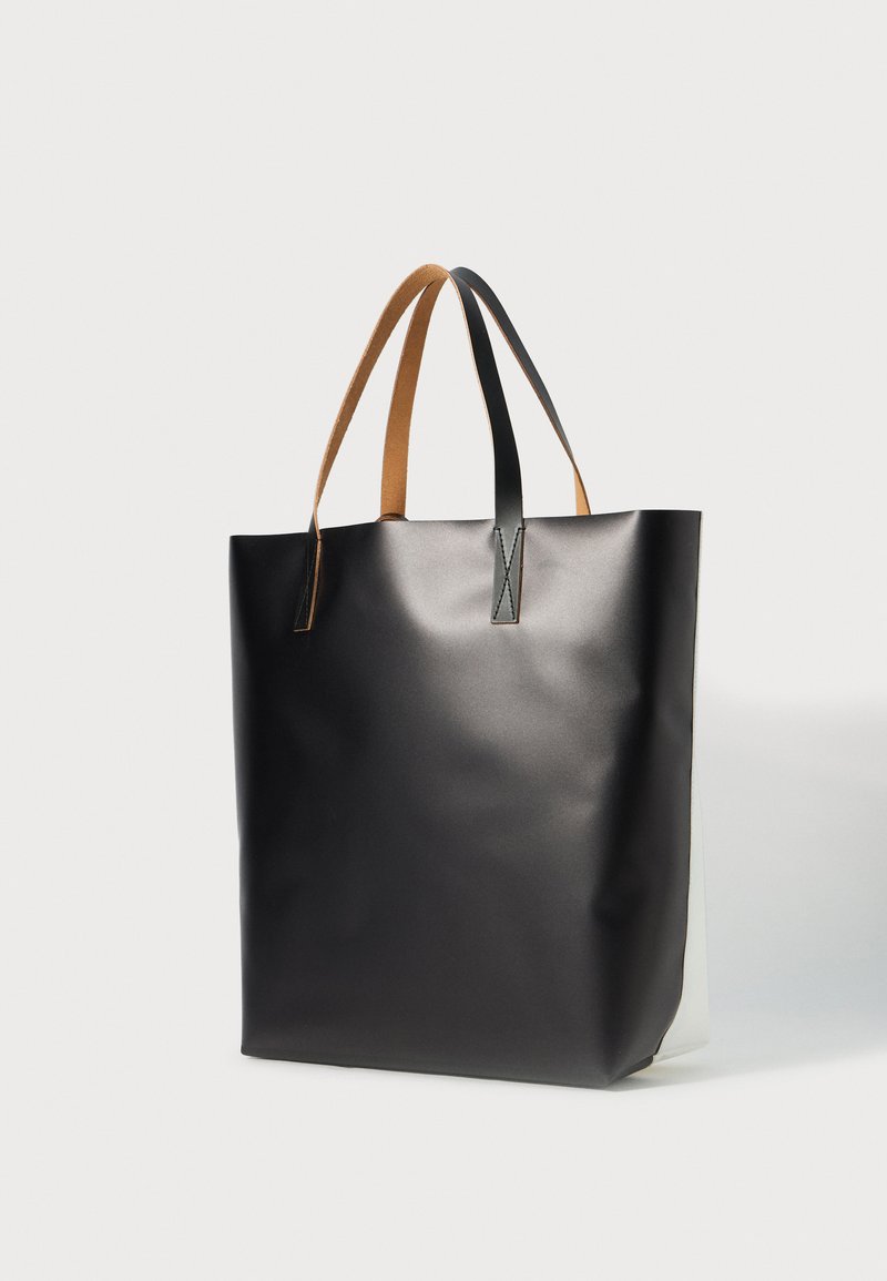 Sac fourre-tout noir avec une surface lisse, des poignées marron et un design minimaliste. Présente une forme plate et un intérieur blanc contrastant.