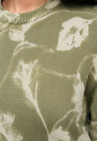 Alyvučių žalia megzta palaidinė, puošta šviesiai kreminu gėlių raštu. Apvalus kaklas ir tekstūruota apdaila suteikia dizainui detalių.