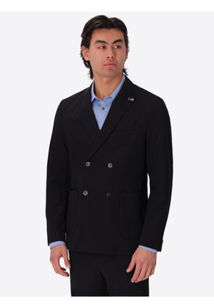 Uomo che indossa un blazer nero doppiopetto sopra una camicia azzurro chiaro con colletto, in piedi con le mani rilassate, che guarda di lato.