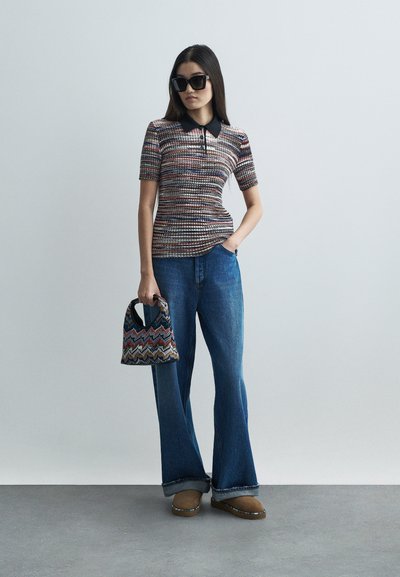 Polo s krátkým rukávem s mnohobarevným tkaným vzorem, černým límečkem, v kombinaci s modrými rozšířenými džíny a vzorovanou kabelkou. Hnědé nazouvací boty.