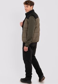 Padded Weste mit olivgrünem Körper und schwarzen Schultern, ausgestattet mit einem hohen Kragen, Reißverschluss und Seitentaschen, getragen über einem langärmligen Hemd.