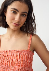 Smocked crop top in bright orange fabric with a textured pattern and thin adjustable straps, featuring a checkered design on the straps.