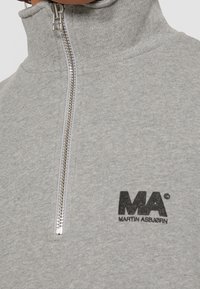 Sudadera gris con cierre de cremallera y cuello alto, con una cremallera plateada y un logo impreso negro "MA" en el pecho izquierdo. Textura suave.