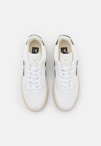 Pár bílých tenisek Veja s černým logem po stranách, béžovou podrážkou, perforovanou špičkou a bílými tkaničkami, pohled zhora na bílém pozadí.