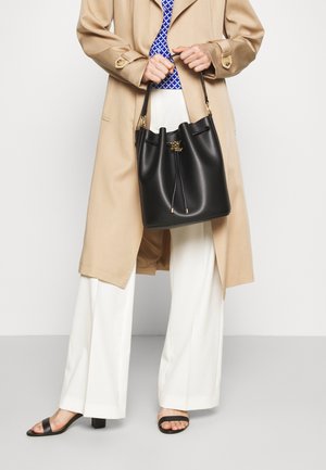 Person iført beige trenchcoat og hvide bukser, der holder en sort læderbucket-taske med gulddetaljer foran en hvid baggrund.
