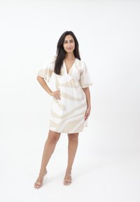 Kurzärmliges, V-Ausschnitt-Kleid in Creme mit beigen abstrakten Mustern. Der Stoff ist glatt. Klare Absatzsandalen ergänzen das Outfit.