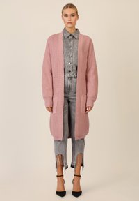 Roze gebreide cardigan met geribbelde textuur, open voorkant en zakken, gedragen over een grijze button-up shirt en lichtgrijze versleten jeans met franjes.